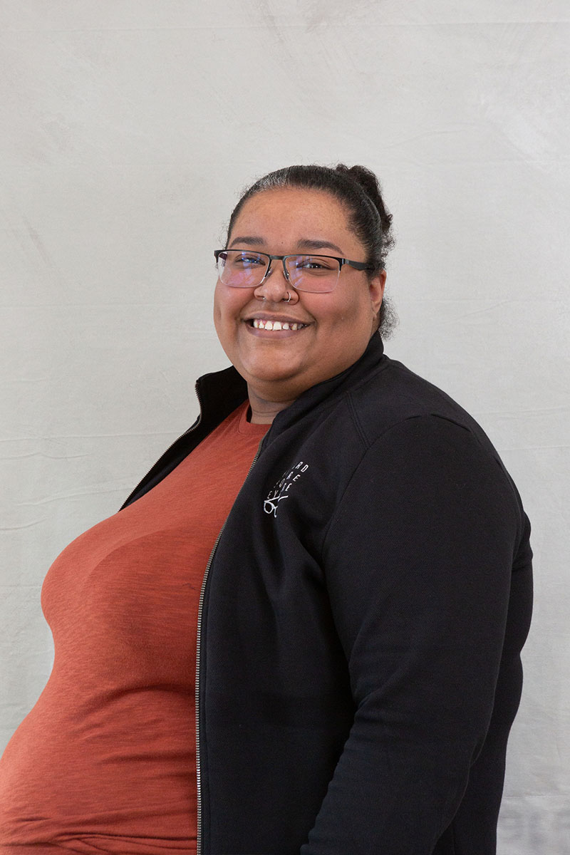 The image depicts a person standing against a white background, smiling at the camera, wearing glasses, a black jacket with a zipper, and a light-colored shirt underneath.