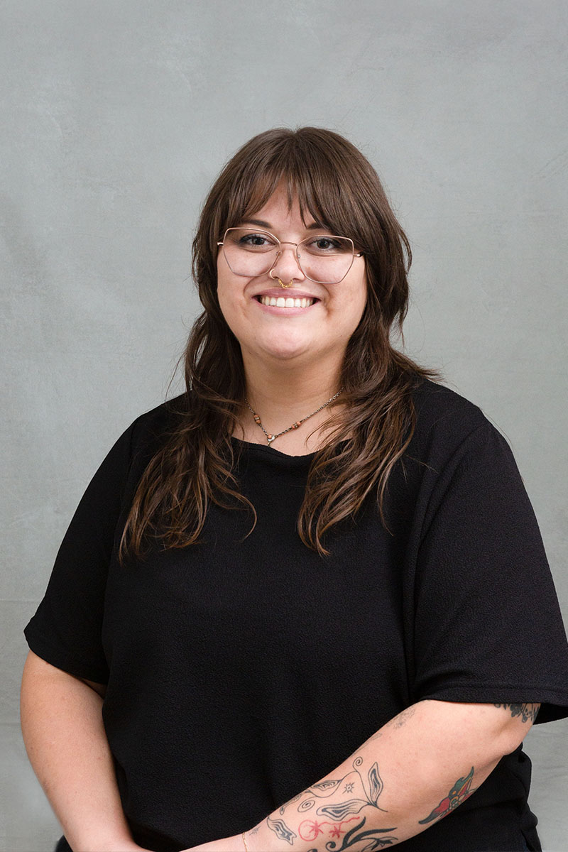 The image depicts a person standing against a light background with a neutral expression, wearing glasses, a black top, and a tattoo on their left arm.