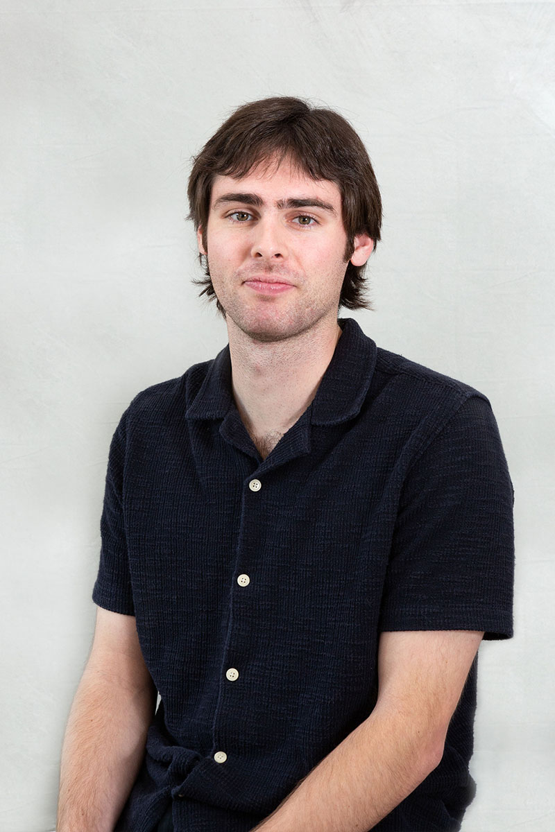 The image shows a man with short hair wearing a dark blue polo shirt and sitting against a plain background.