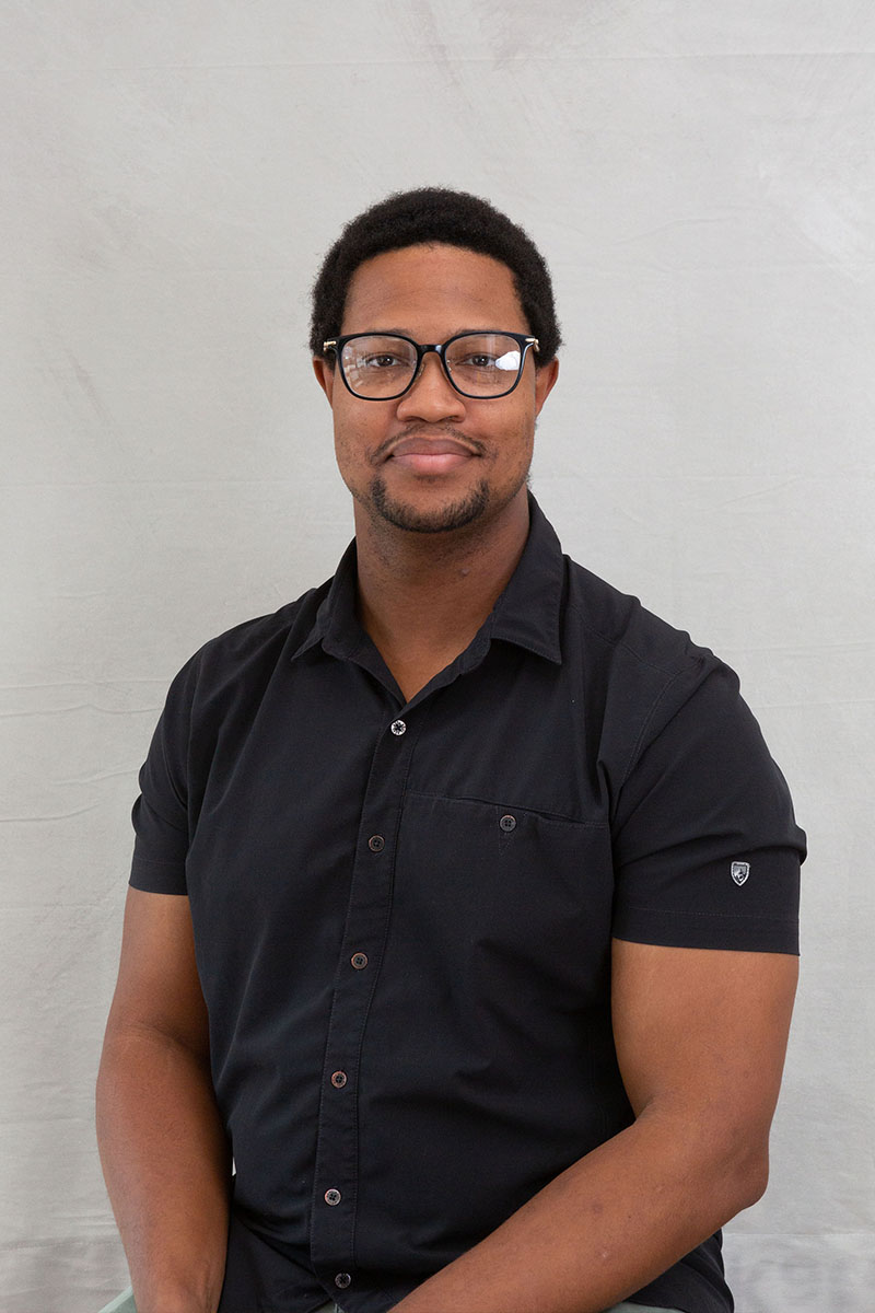 The image shows a man sitting with his arms crossed, wearing glasses, a dark shirt, and a black vest, against a white background.