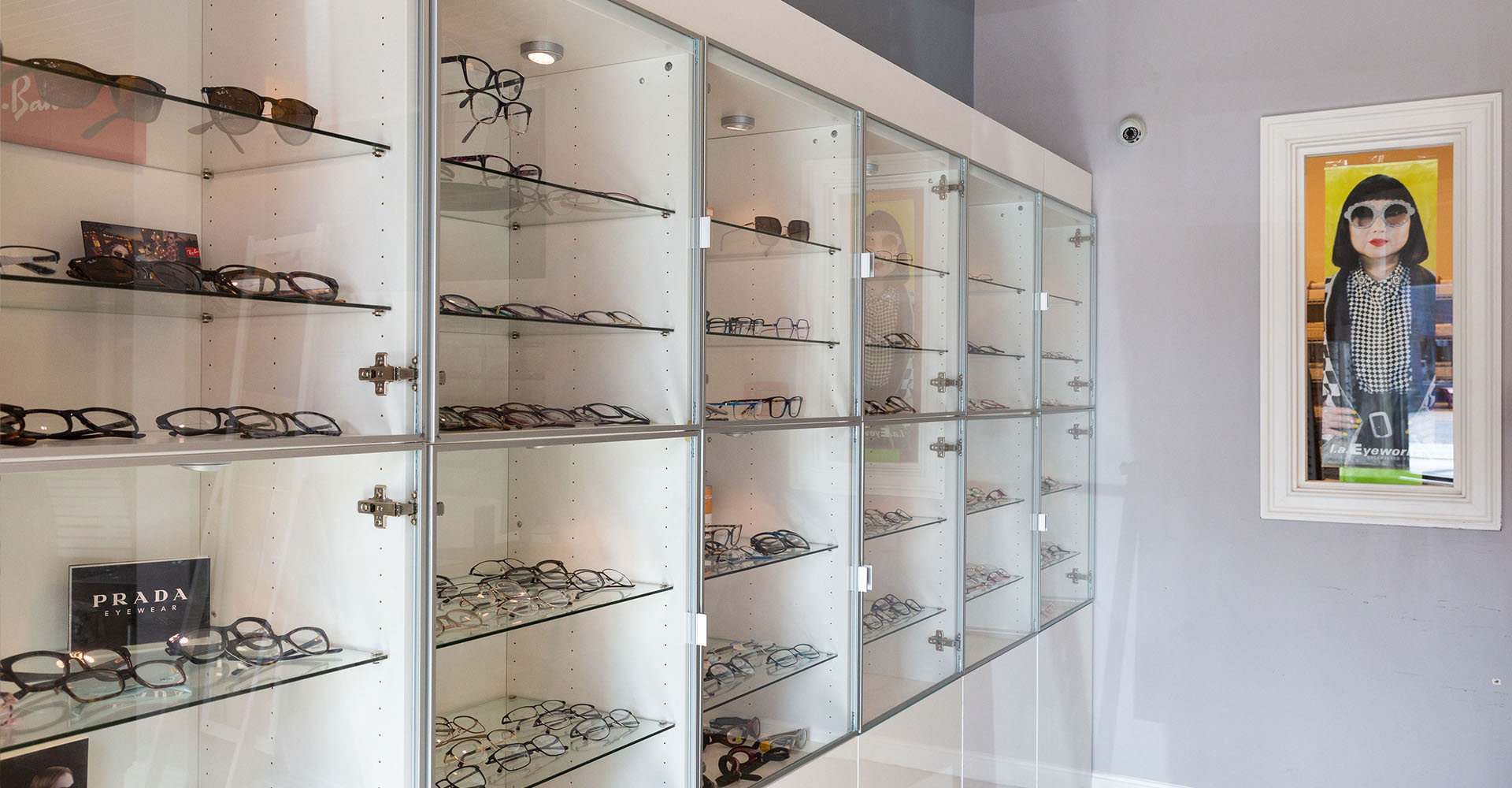The image shows a display case filled with various styles of eyeglasses, with a framed picture hanging above it.