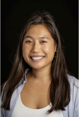 The image shows a person smiling at the camera, with short hair, wearing a blue top and standing against a dark background.