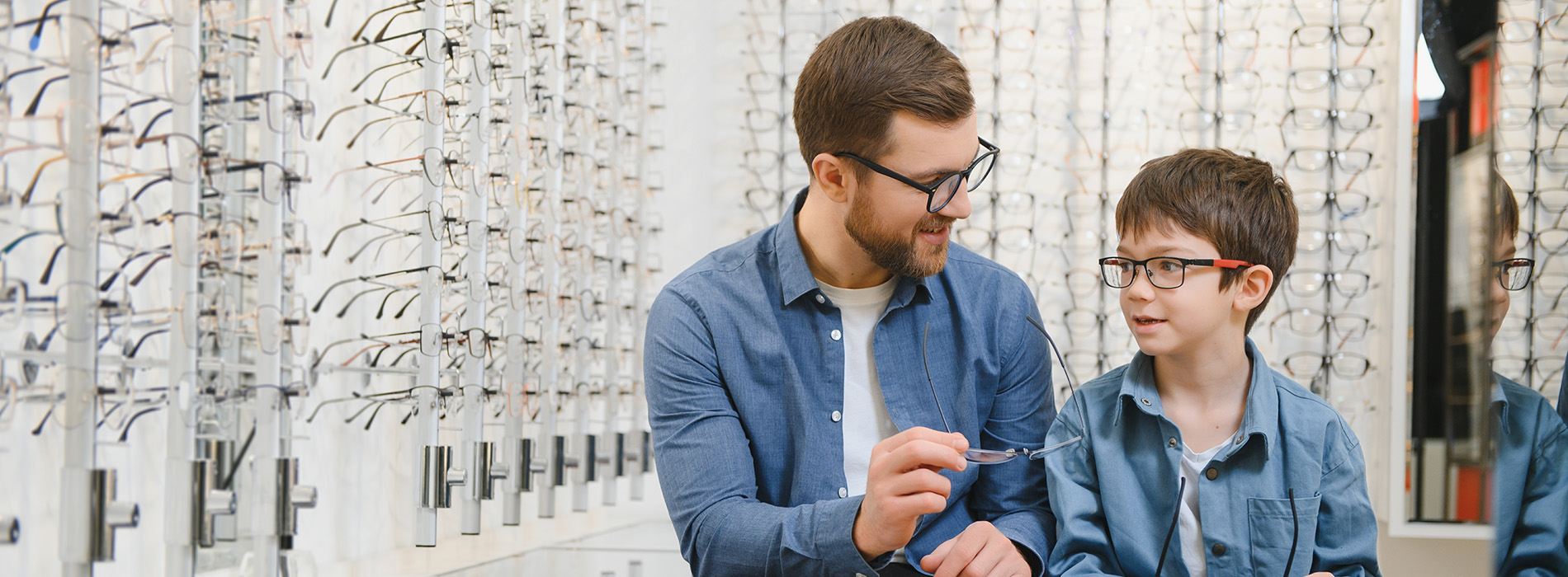 A man and a young boy are standing in front of an optical display with various frames on shelves behind them. The man appears to be showing something to the boy, possibly explaining eyeglasses.