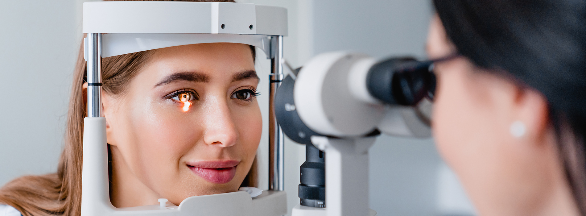 The image shows a woman sitting in front of an eye machine with her eyes closed, while another person is standing behind her, looking at the screen of the machine.