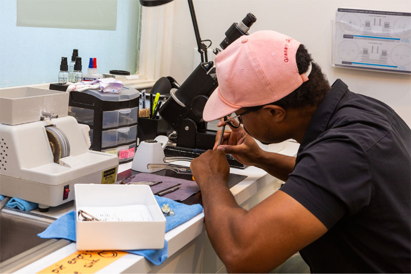 This is a photograph of a person working at a microscope, likely preparing samples or conducting scientific research, with various lab equipment visible in the background.
