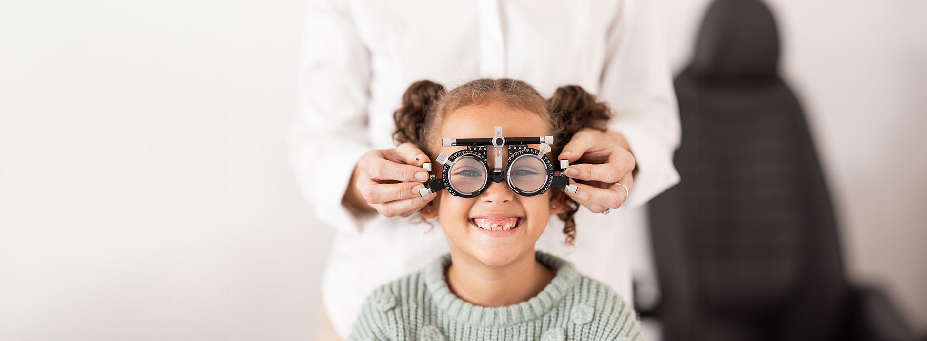 A young child in front of an adult holding up a pair of glasses, both indoors with a blurred background.