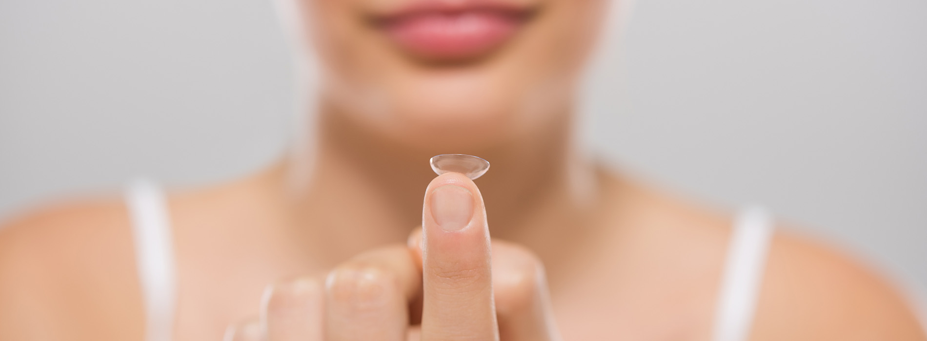 The image shows a close-up of a person s hand holding a contact lens with a visible reflection of a face in the center, set against a blurred background that appears to be a human head.