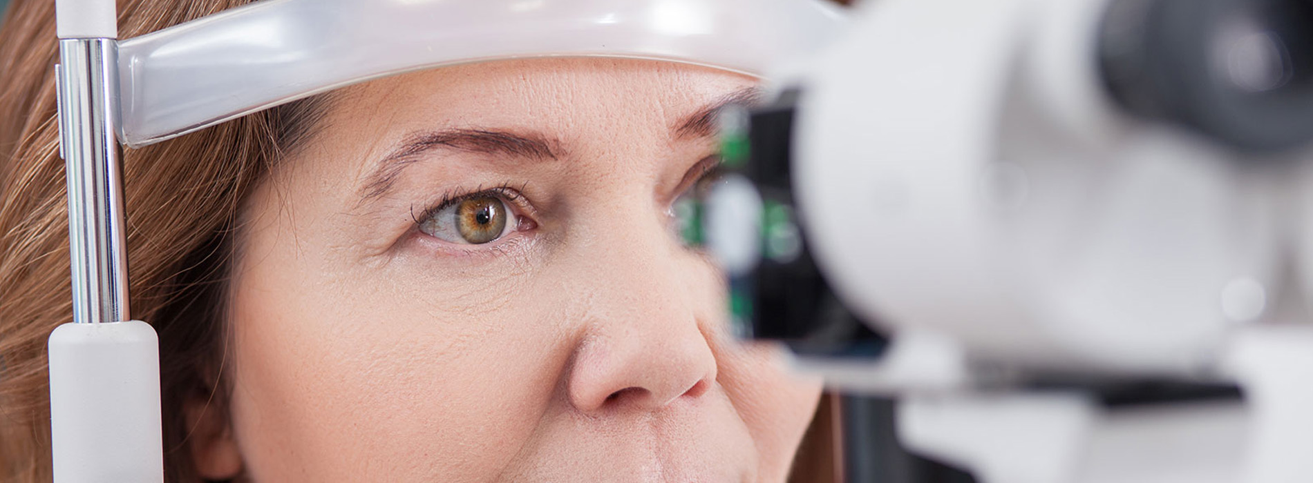 A woman with brown hair is seated at an eye examination machine, looking into a device with her face partially obscured by the equipment s frame.