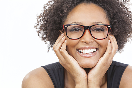 A woman with curly hair, wearing glasses, smiling broadly at the camera with her hands on her cheeks.