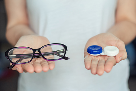 The image shows two hands holding a blue pill and a pair of glasses, with a backdrop of a person s arm and shoulder.